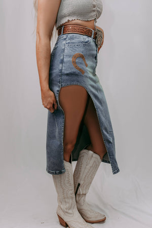 Person wearing a denim skirt with a horse design and white boots on a plain background