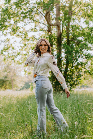Woman in a white blouse with floral patterns and light blue jeans standing in a grassy field with trees in the background.