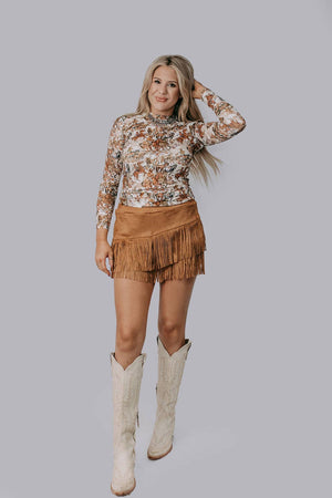 Woman wearing a floral top and brown fringed skirt with white boots on a gray background