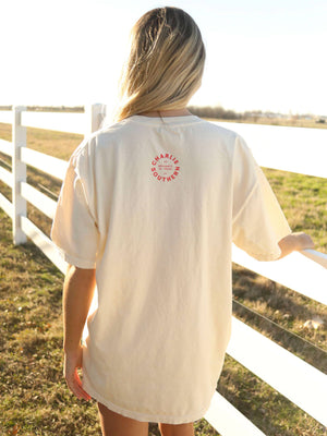 Person wearing a cream-colored t-shirt with a logo, standing in a field with a white fence.