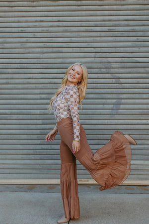Woman in a floral top and brown bell-bottom pants posing against a metal shutter.