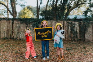 three kids holding a rectangular black flag with the phrase 'Raise Hell, Kid' screen-printed in gold lettering, with gold details along the edges.
