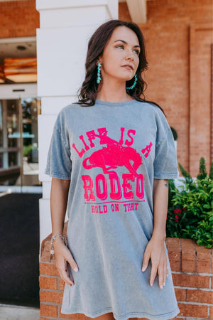 Woman wearing a gray t-shirt with pink 'Life is Rodeo' design in front of a brick building.