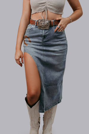 Person wearing a denim skirt with a slit, beige top, and boots on a plain background