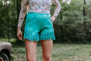 Person wearing a floral top and teal fringed shorts in a natural setting