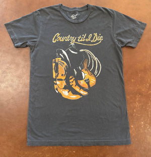Gray t-shirt with a graphic of cowboy boots, guitar, and text 'Country til I Die' on a brown surface.