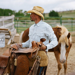 Man in cowboy hat with a horse and saddle in an outdoor setting