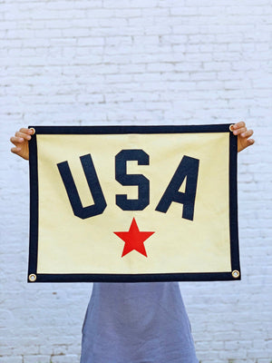 A rectangular camp flag with a cream body, featuring the letters 'USA' in navy blue and a red star below, bordered with a navy detail.