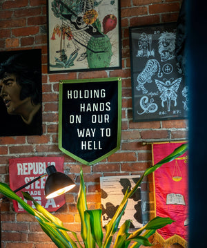 Wall art on a brick wall. The main one is a pennant that says "Holding Hands on our way to Hell". 