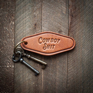 Leather keychain with 'Cowboy Shit' tag on a wooden surface
