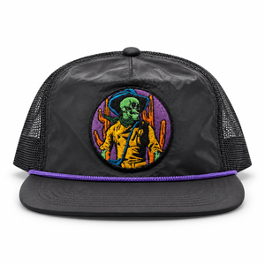 A black trucker hat with a colorful embroidered circular patch on the front. The patch features a green-skinned alien in a yellow jacket surrounded by purple and orange desert cacti.