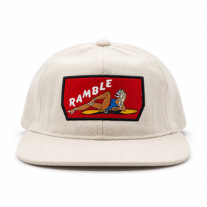 An off-white baseball wool snapback cap with a red rectangular embroidered patch on the front featuring the word 'RAMBLE' in white and a cartoon illustration of a woman on a plane propeller. 