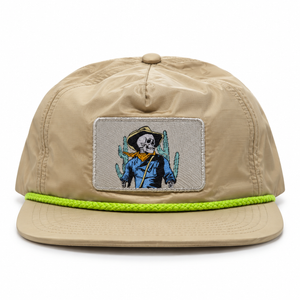 A beige 5-panel cap with a flat brim, featuring a bright neon green braided trim around the edge of the brim. The front panel has a rectangular patch depicting a skeleton cowboy wearing a blue shirt and hat with a yellow bandana, standing in front of green cacti.