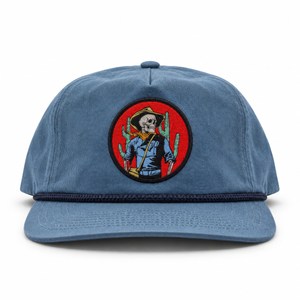 A blue cotton twill snapback cap featuring a circular woven patch on the front with a skeleton cowboy holding a cactus against a red background.