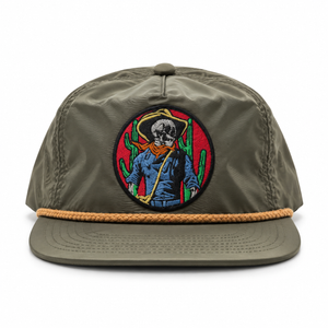 An olive green flat-brim cap with a tan braided rope detail around the brim and a circular embroidered patch on the front featuring a skeleton cowboy with a cactus and red background.
