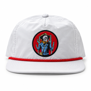A white flat-brim cap with a red braided trim along the edge of the brim and a circular woven patch on the front featuring a skeleton soldier with cactus details.