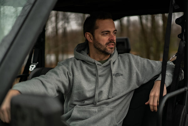 Man sitting inside a vehicle wearing a gray hoodie with a logo, surrounded by a forest.
