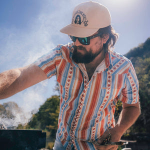 Man wearing a patterned shirt and cap with sunglasses, standing outdoors.