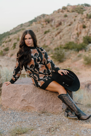 Woman in a patterned dress and cowboy boots sitting on a rock with a mountainous background